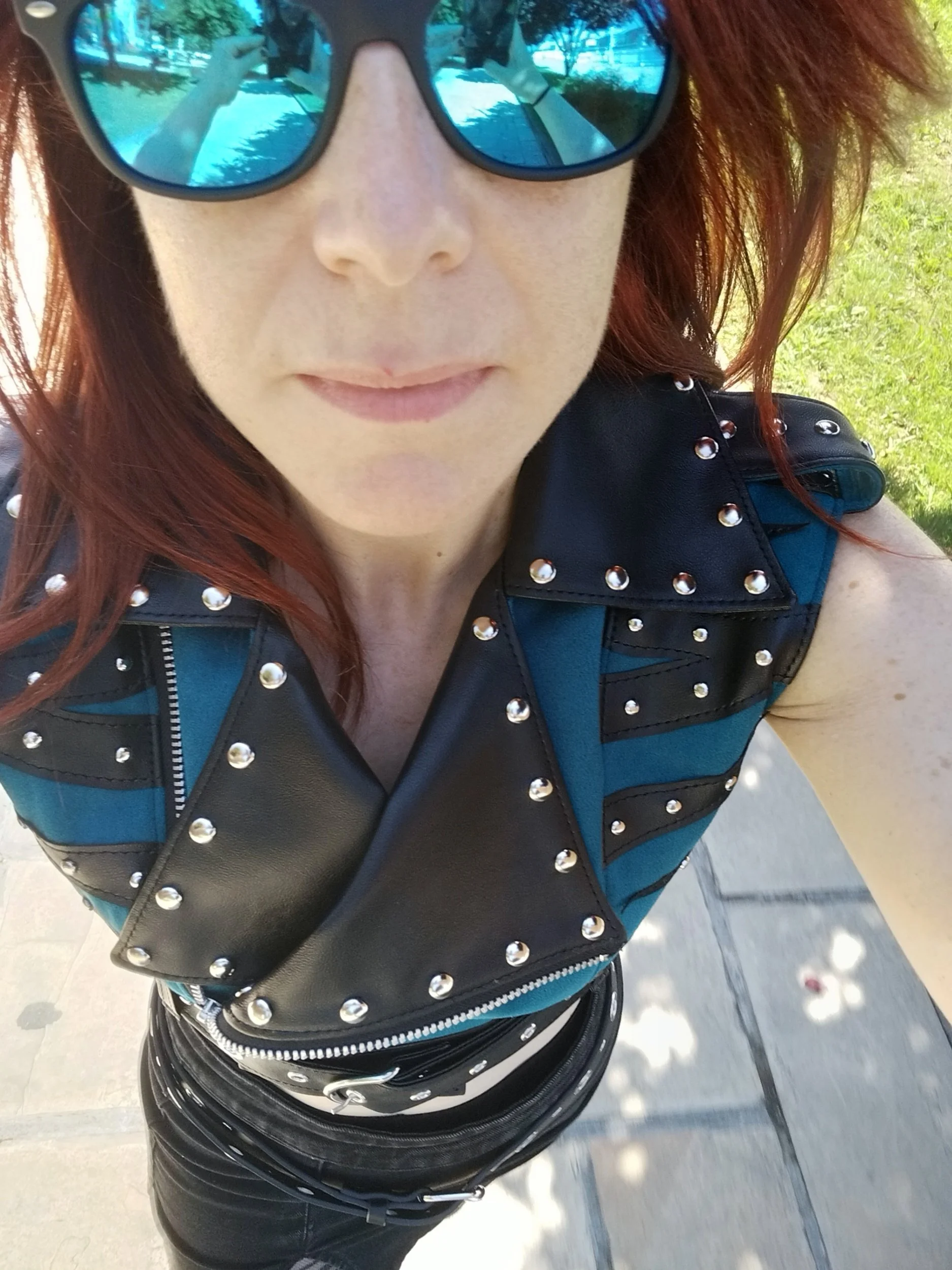 Close-up of a woman with red hair wearing black sunglasses, a black leather vest with silver studs, and a belt with silver studs, taking a selfie outdoors on a sunny day.