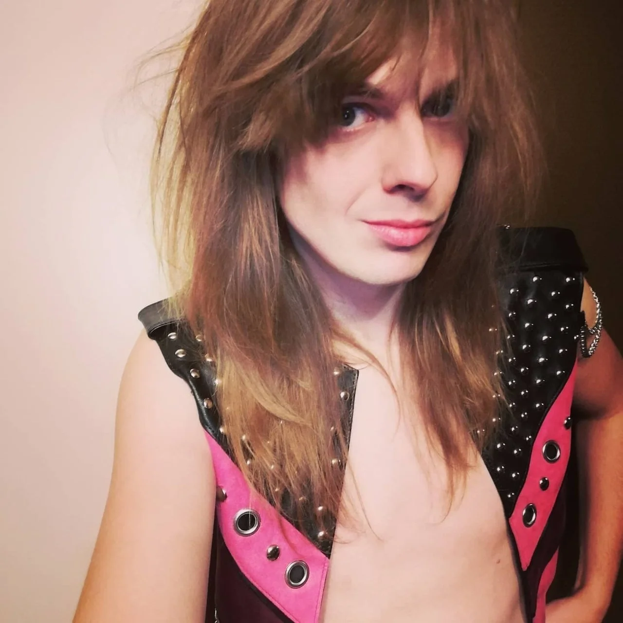 A person with long, wavy, light brown hair wearing a black and pink sleeveless vest with silver studs and eyelets, posing against a neutral background.