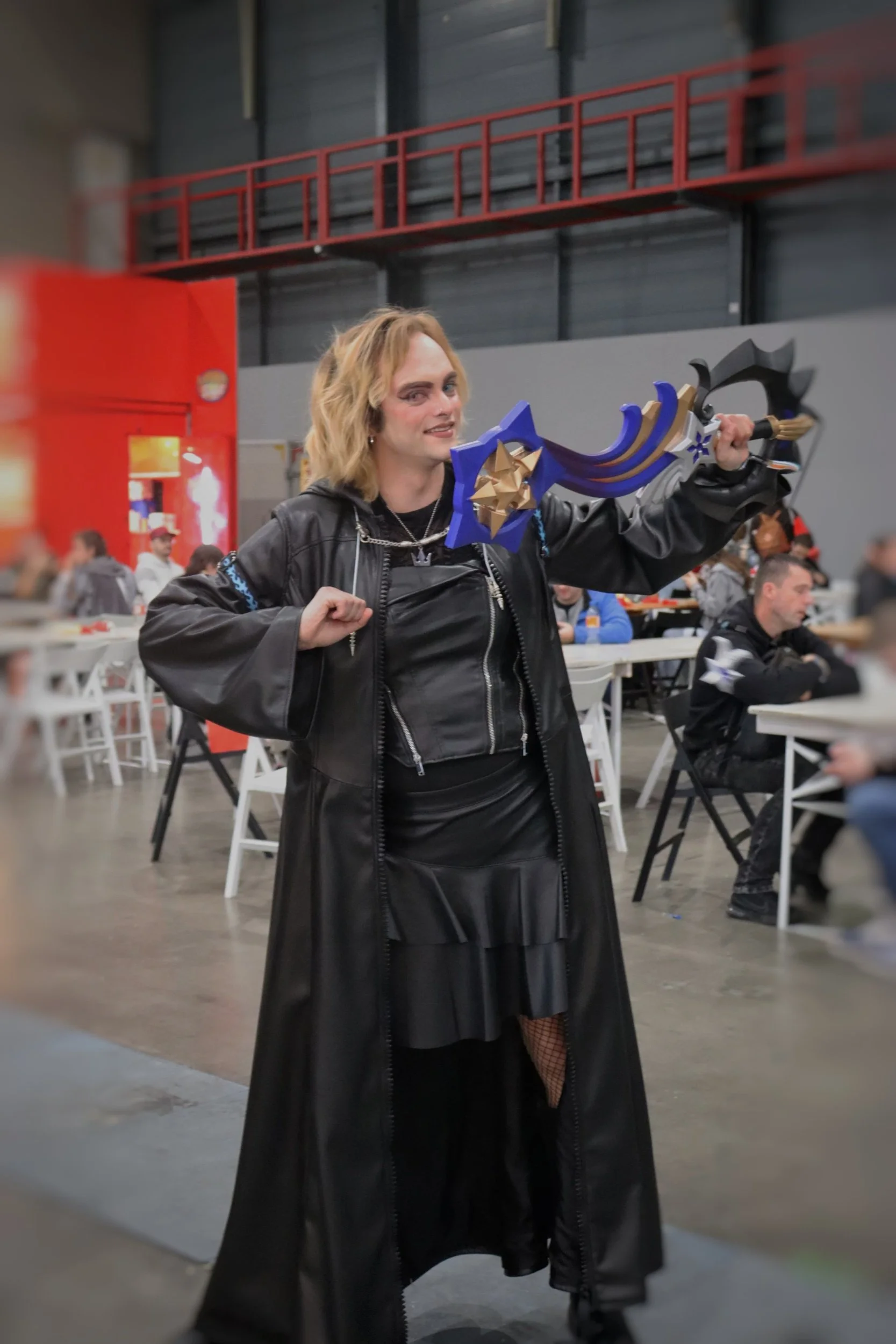 Person in cosplay costume holding a large, decorative sword, at a convention or event with tables and other attendees in the background.