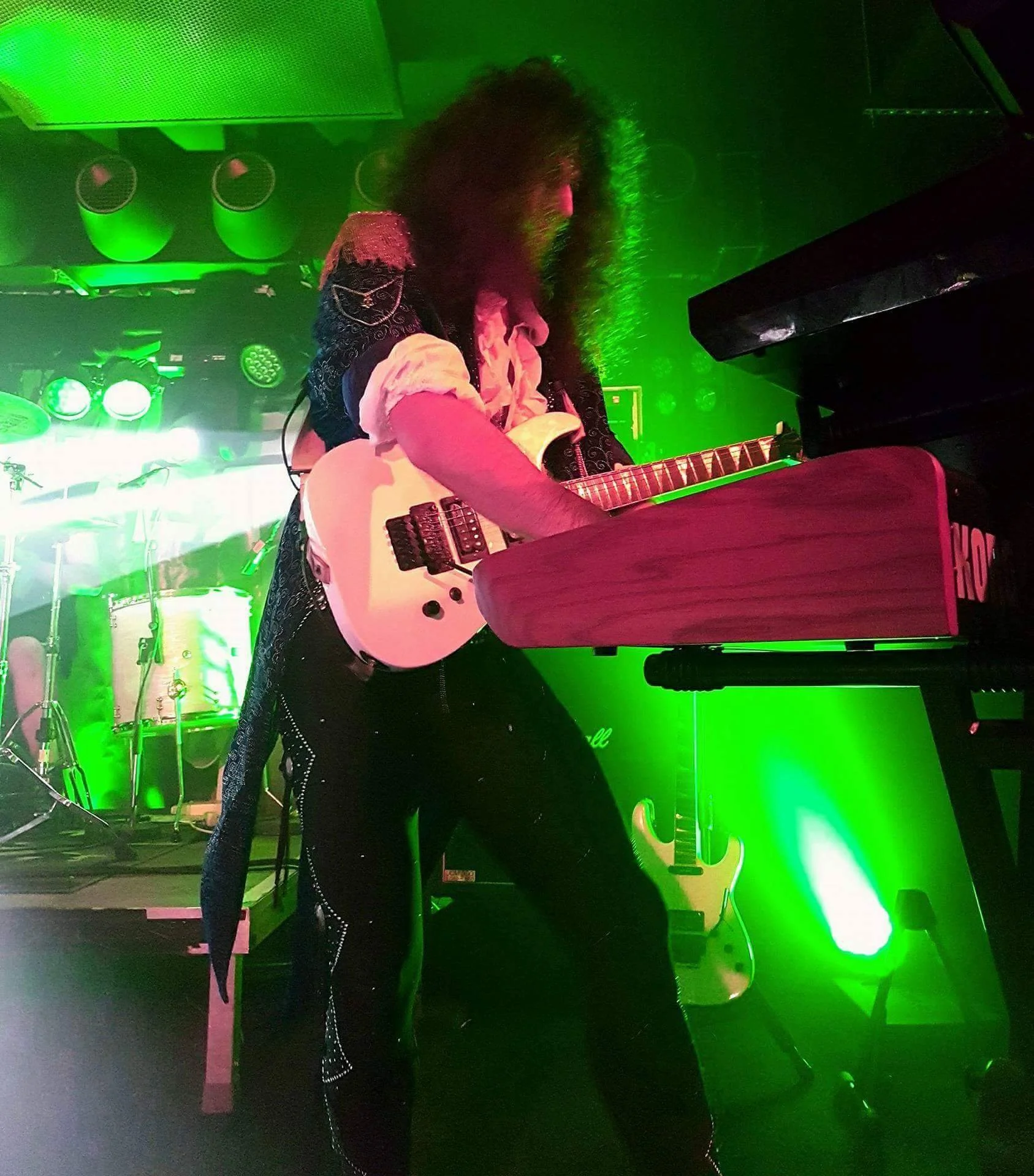 Person playing guitar and keyboard on stage with green lighting, wearing dark clothing with decorative details, curly hair, and a microphone in front.