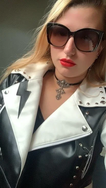 A woman with blonde hair wearing large black sunglasses, red lipstick, a black choker with a silver pendant, and a white and black studded leather jacket with a lightning bolt design.
