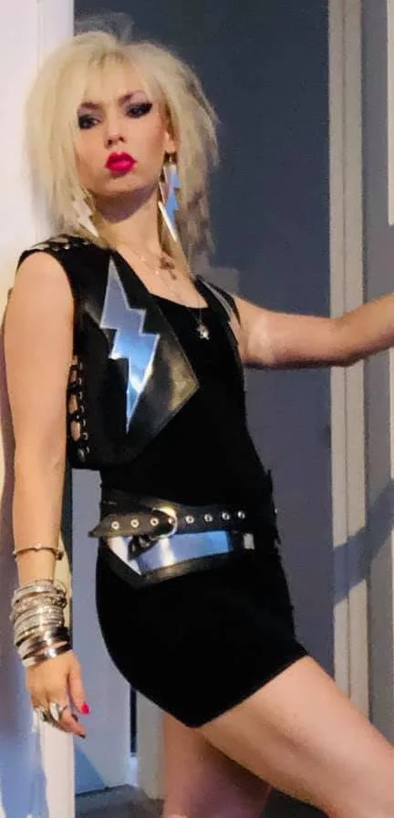 A woman with platinum blonde, messy hair and bold makeup, wearing a black dress with lightning bolt patches, multiple bracelets, and a thick belt, striking a confident pose.