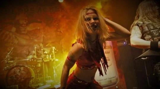 A female singer with long hair and tattoos performing on stage with bright yellow and orange lights, and a drummer visible in the background.
