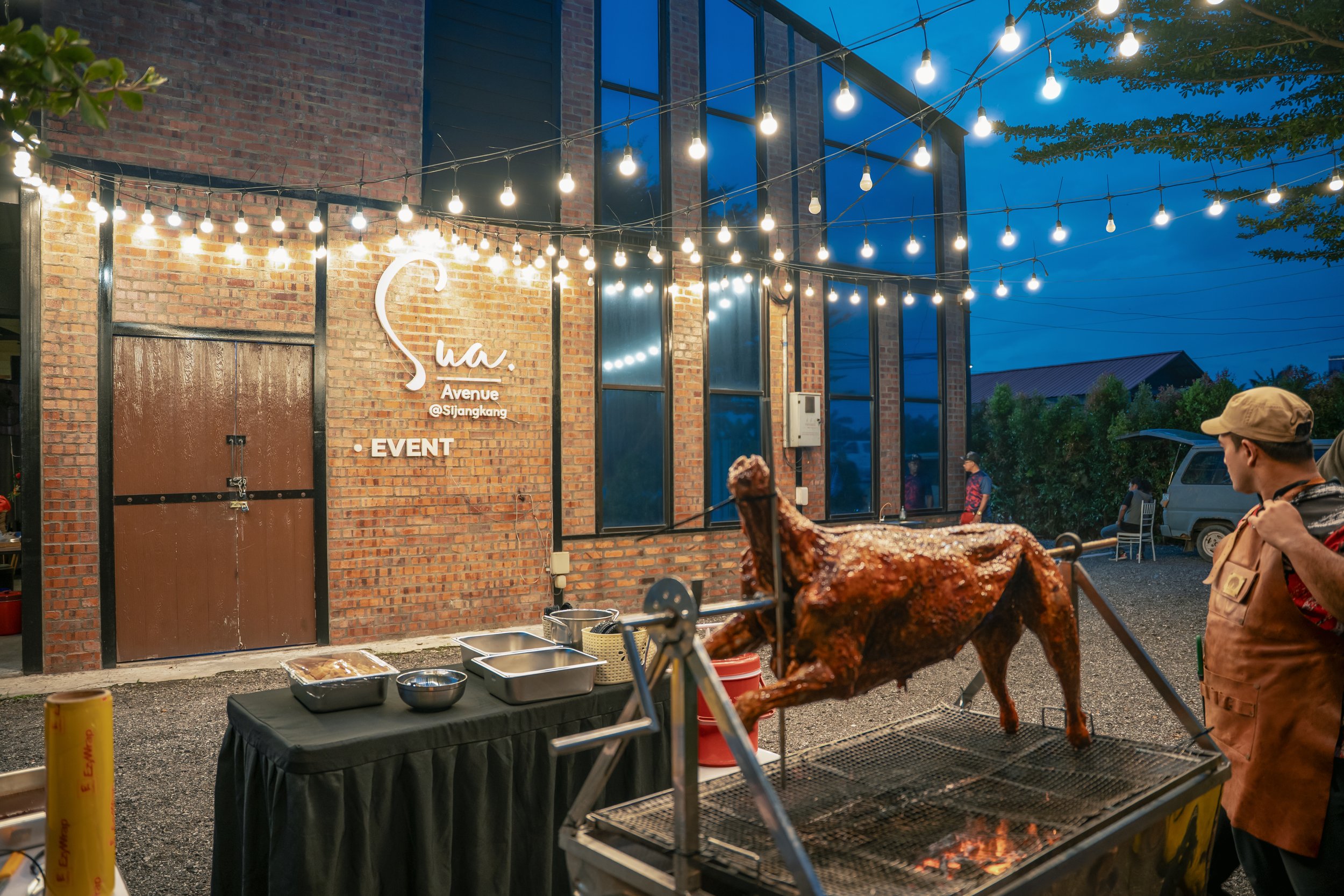 An outdoor evening scene with a roasted whole pig on a spit over a grill, a man wearing a cap and apron standing nearby, and a brick building decorated with string lights. The brick building has a sign that reads '@Sijangkang' and 'EVENT', with large windows and a door.
