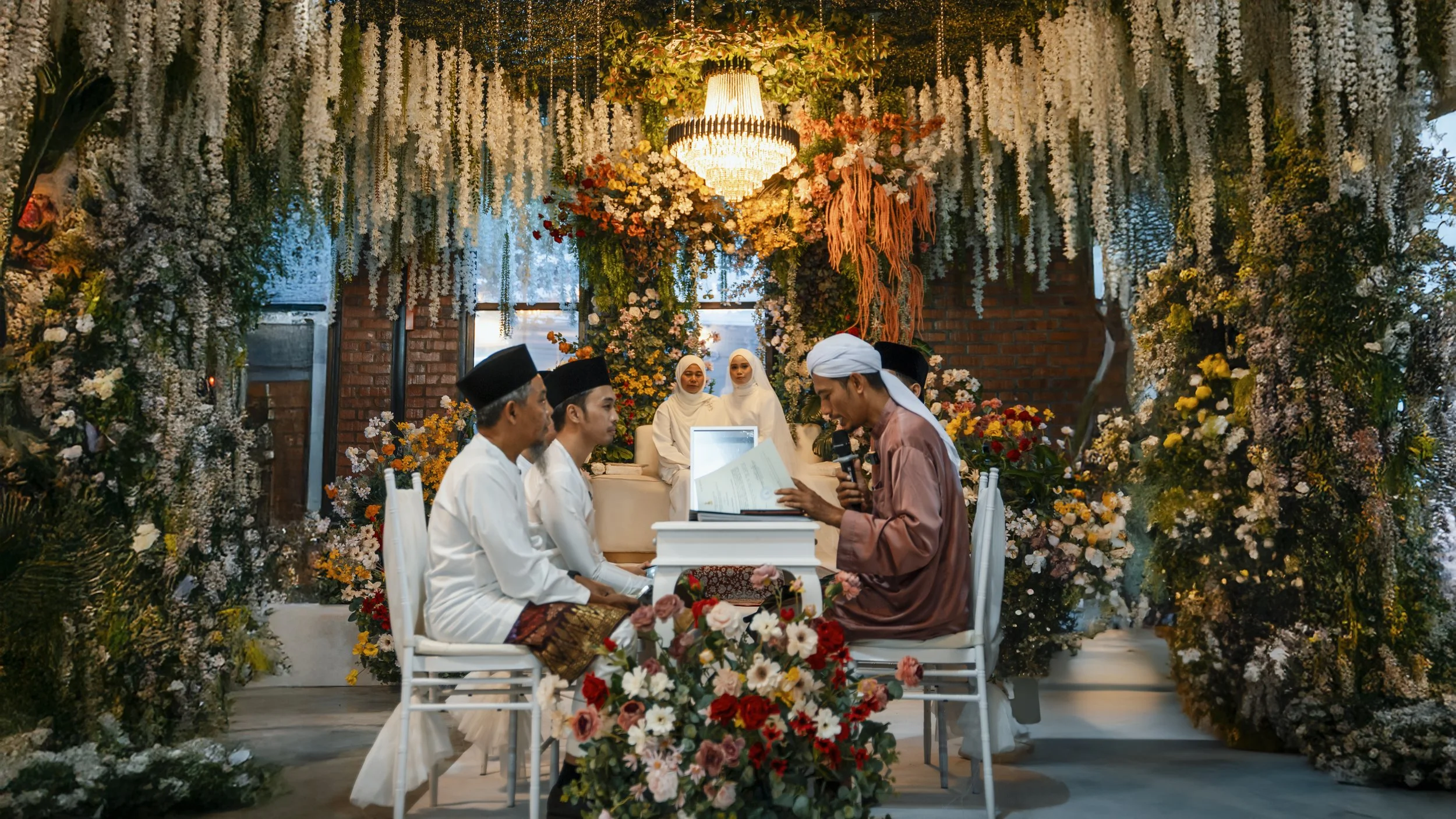 A traditional Muslim wedding ceremony with five individuals dressed in Islamic attire, seated at a table surrounded by elaborate floral decorations, with a bride and groom in white and light-colored clothing, and two women in modest white dresses in the background.