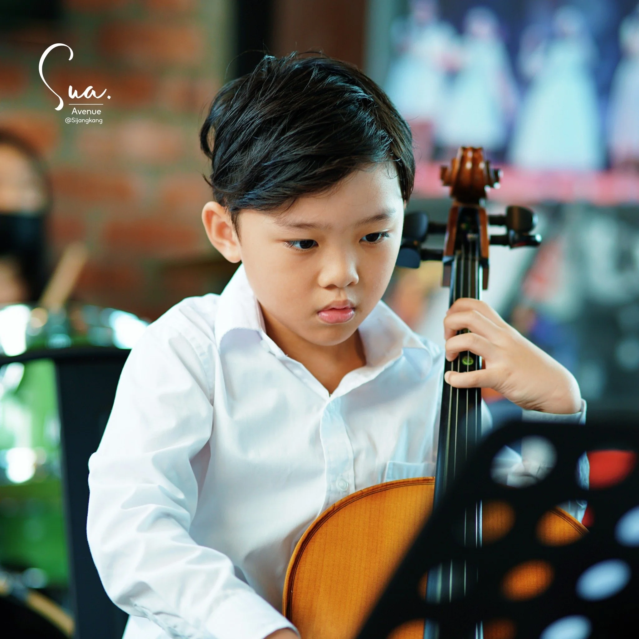 A young boy with black hair wearing a white shirt, playing a violin during a music class or practice session.