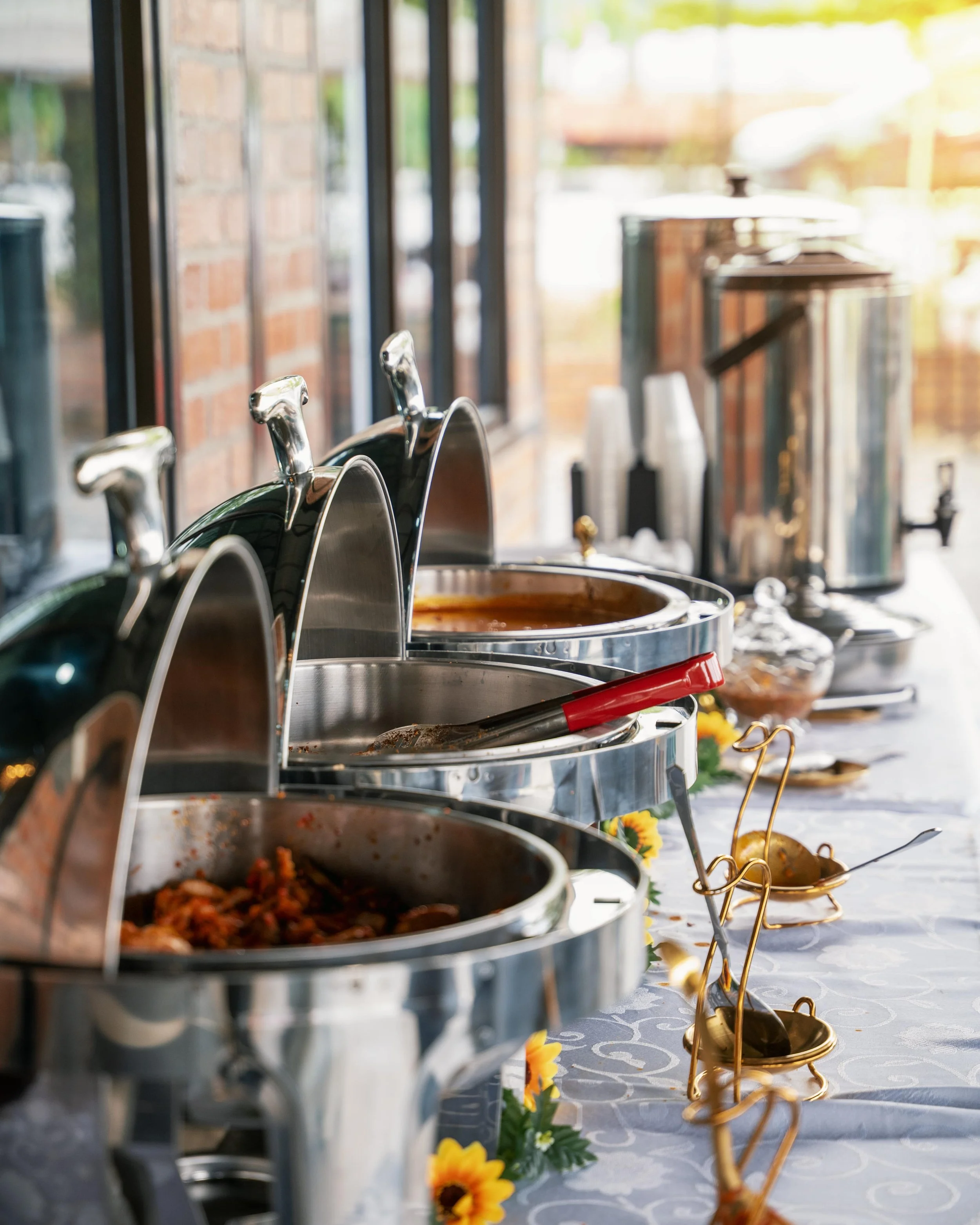 Buffet table with chafing dishes, serving utensils, and decoration, set against a brick wall with a window.