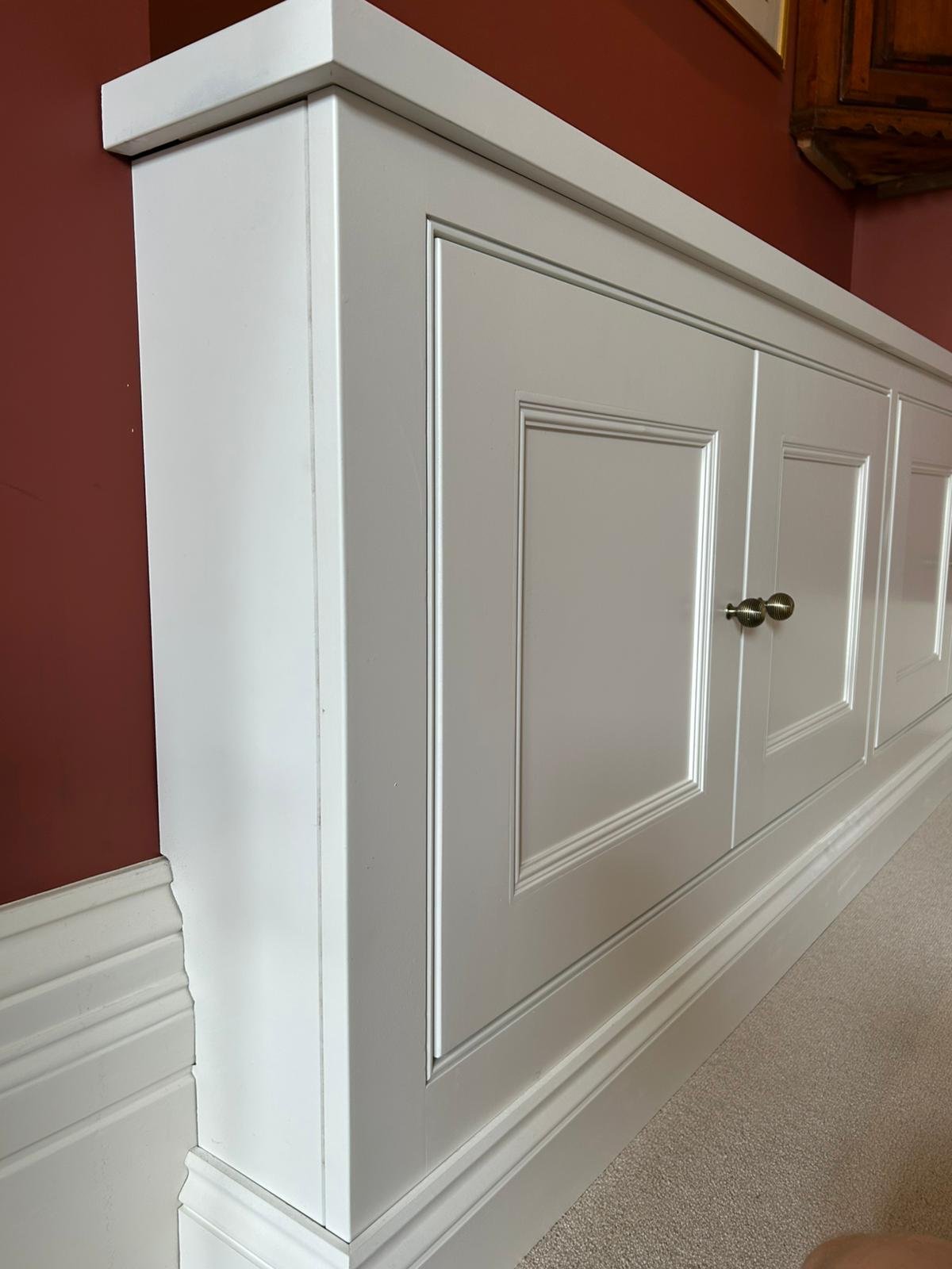 Close-up of a white built-in cabinet with paneled doors and round metallic knobs against a red wall.
