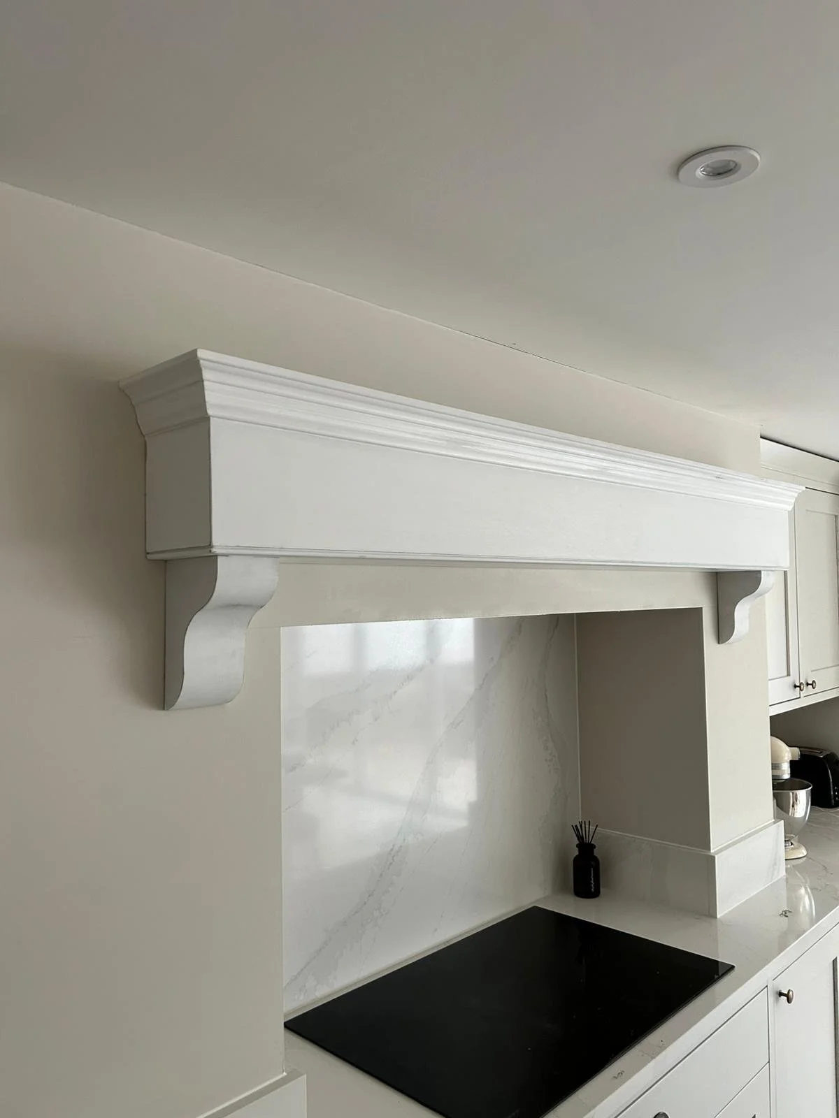 White kitchen shelf with decorative brackets on beige wall above stove, with white cabinets and kitchen appliances in the background.