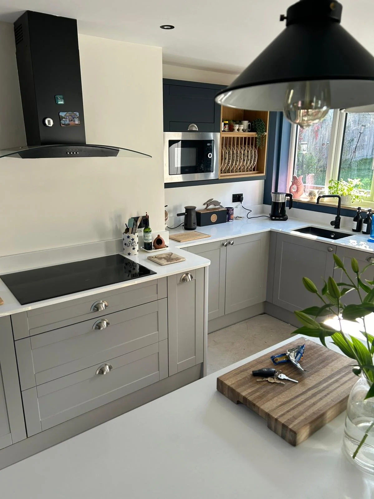 Modern kitchen with light grey cabinets, black stovetop, stainless steel microwave, black range hood, black coffee maker, and a window with a view outside. A wooden cutting board with keys and snacks on a white countertop, and a vase with green plant