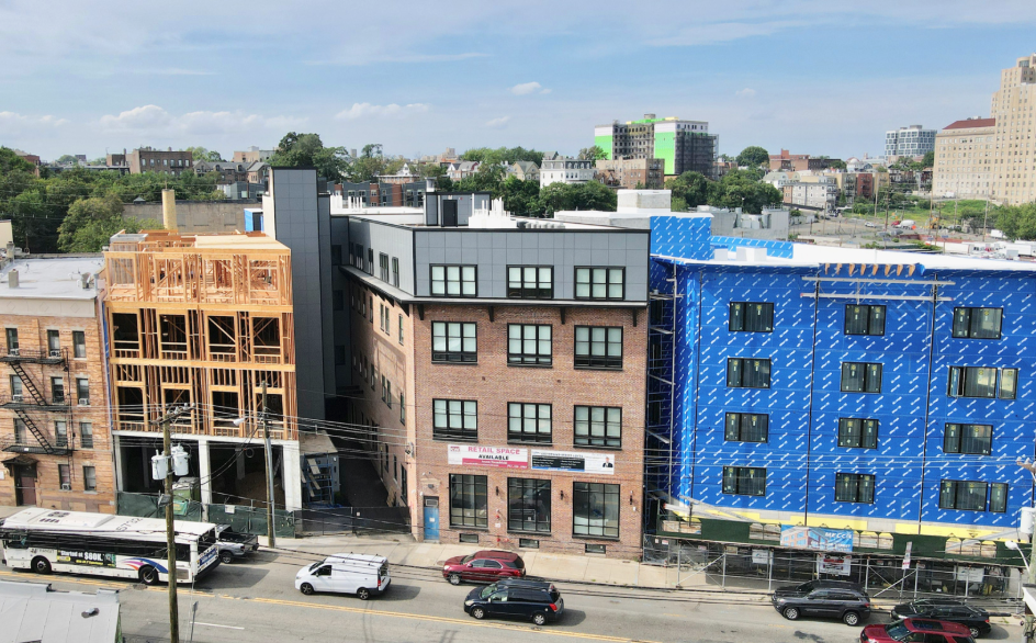 City street view showing buildings under construction, parked cars, and a bus, with a blue building with window coverings, a brick building, and a wooden frame building, under a blue sky with clouds.