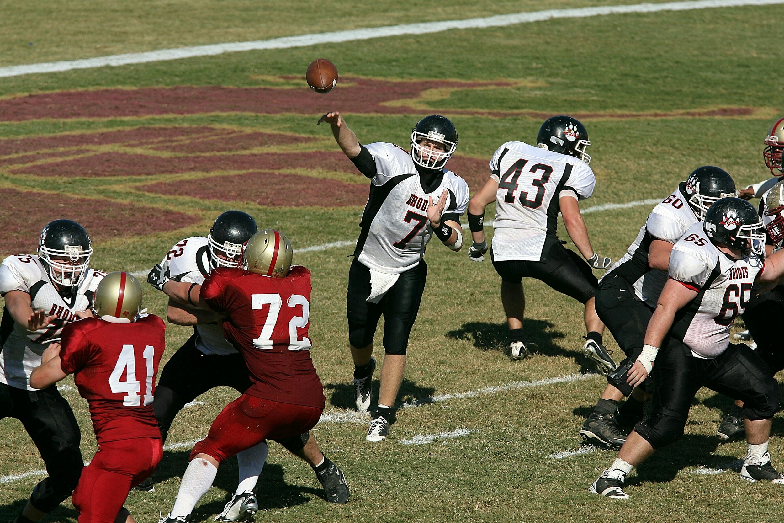 A football game with players in black and white uniforms on offense and players in red and gold uniforms on defense, with one player throwing a pass.