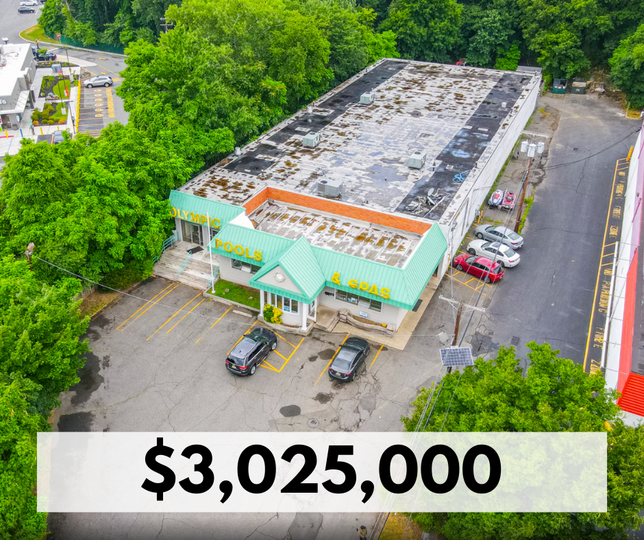Aerial view of a small shopping center with a sign that reads 'Olympic Pools & Spas,' surrounded by parking lots and green trees.