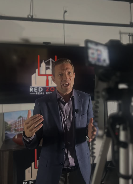 A man in a suit and dress shirt is speaking in front of a camera during a recording or interview in an office setting. Behind him is a screen displaying a logo with a bridge and the text 'RED ZO REAL EST' and a cityscape image.