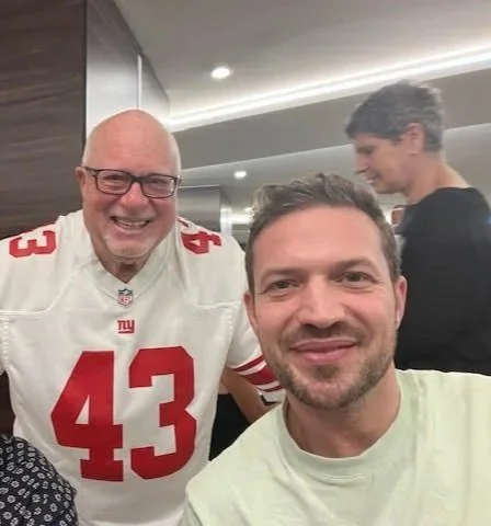 Two men smiling for a selfie, one wearing an NY Giants football jersey number 43, and the other wearing a light-colored shirt, with a woman in the background in a restaurant or cafe setting.