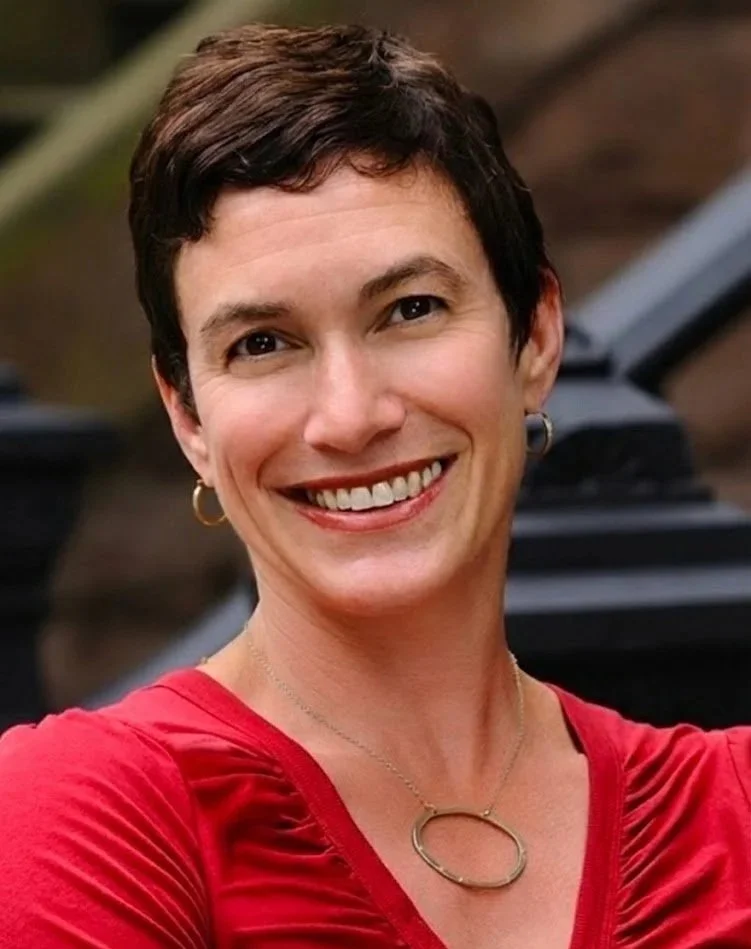 A woman with short dark hair, smiling and wearing a red shirt, with a necklace and earrings, outdoors.