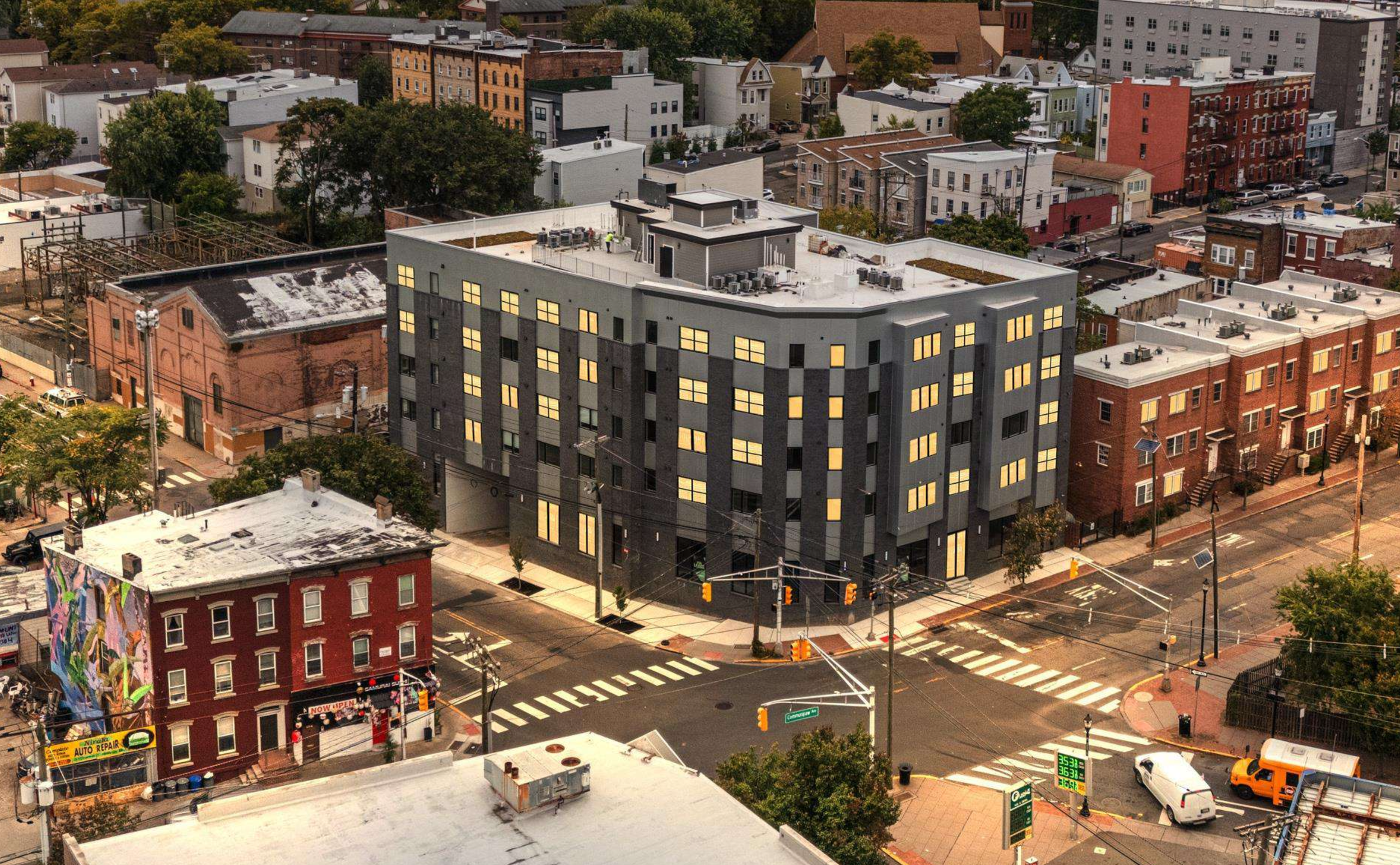 Aerial view of an urban neighborhood with a modern black and gray mid-rise building at an intersection surrounded by older brick and painted buildings, with cars and streetlights.