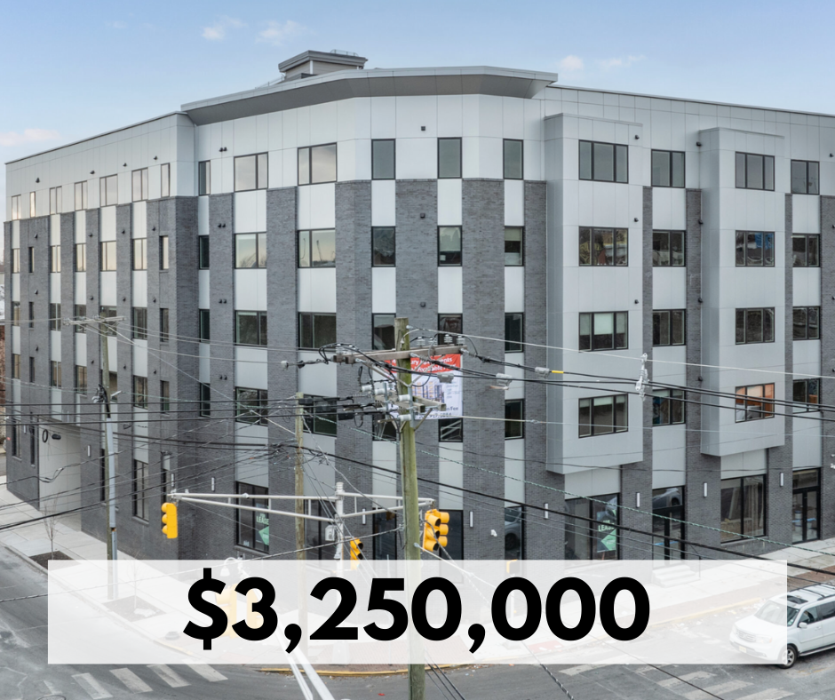 A modern multi-story apartment building with a mix of gray and white exterior, multiple windows, and a corner location, with street traffic, utility poles, and wires in the foreground.