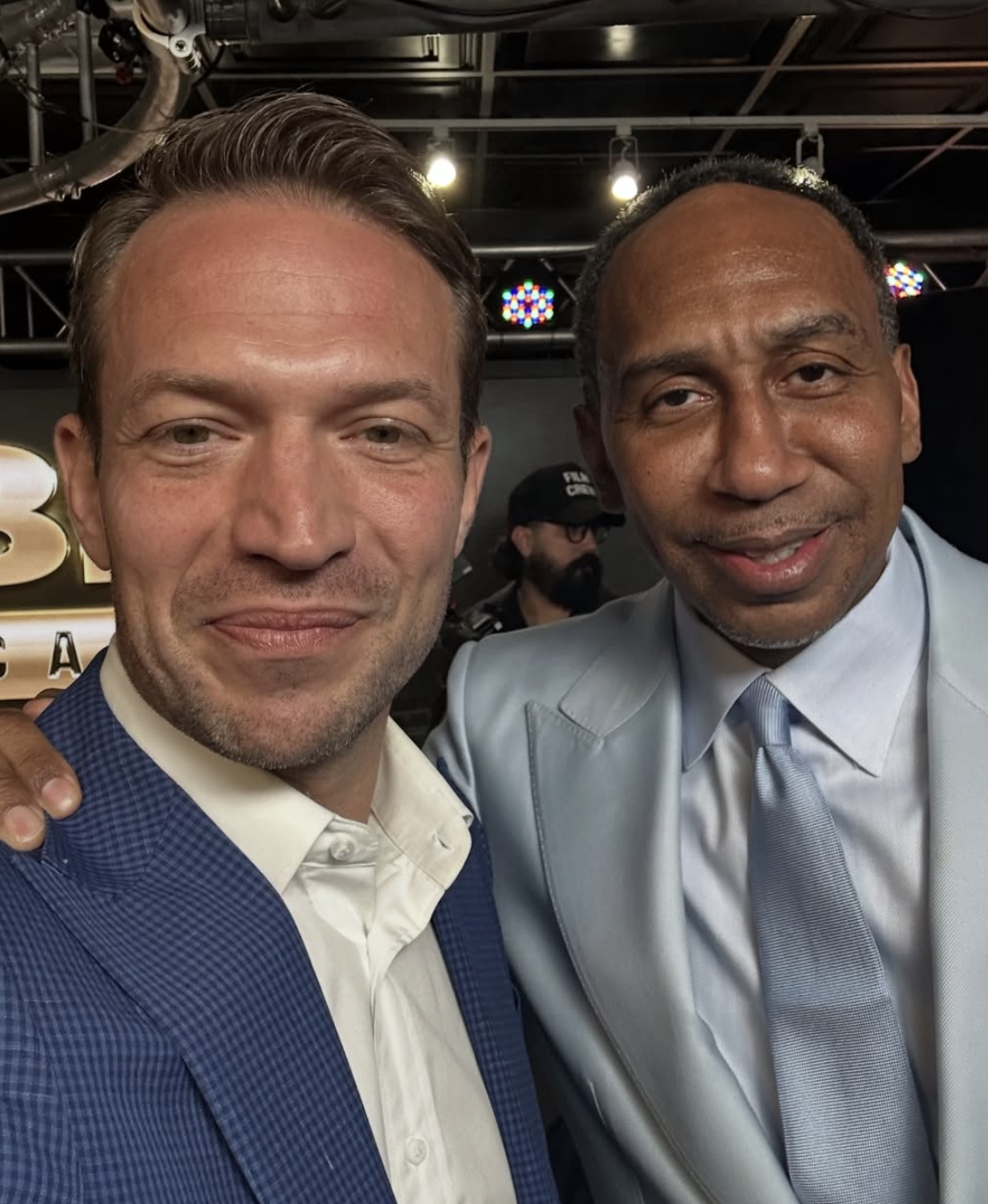 Two men in formal attire taking a selfie at an indoor event, with a person in the background wearing a cap and glasses.