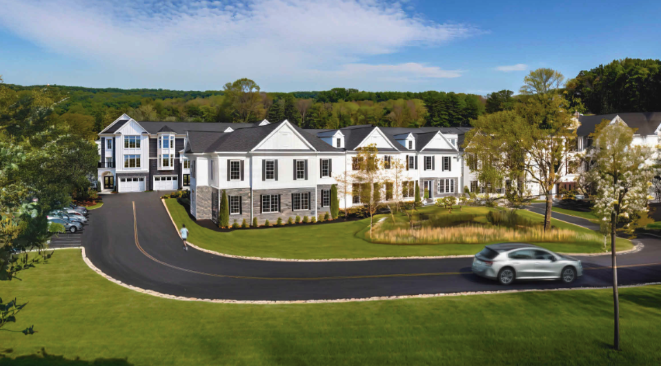 Large white multi-story residential building with black roofs, surrounded by green trees and a well-maintained lawn, with a curved driveway and a silver car driving on it.