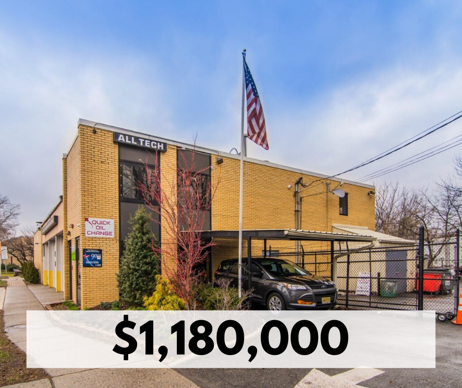A two-story building with a yellow brick exterior, signage for 'All Tech' and 'Quick Oil Change', American flag on a pole, parked car, and surrounding trees and fencing.