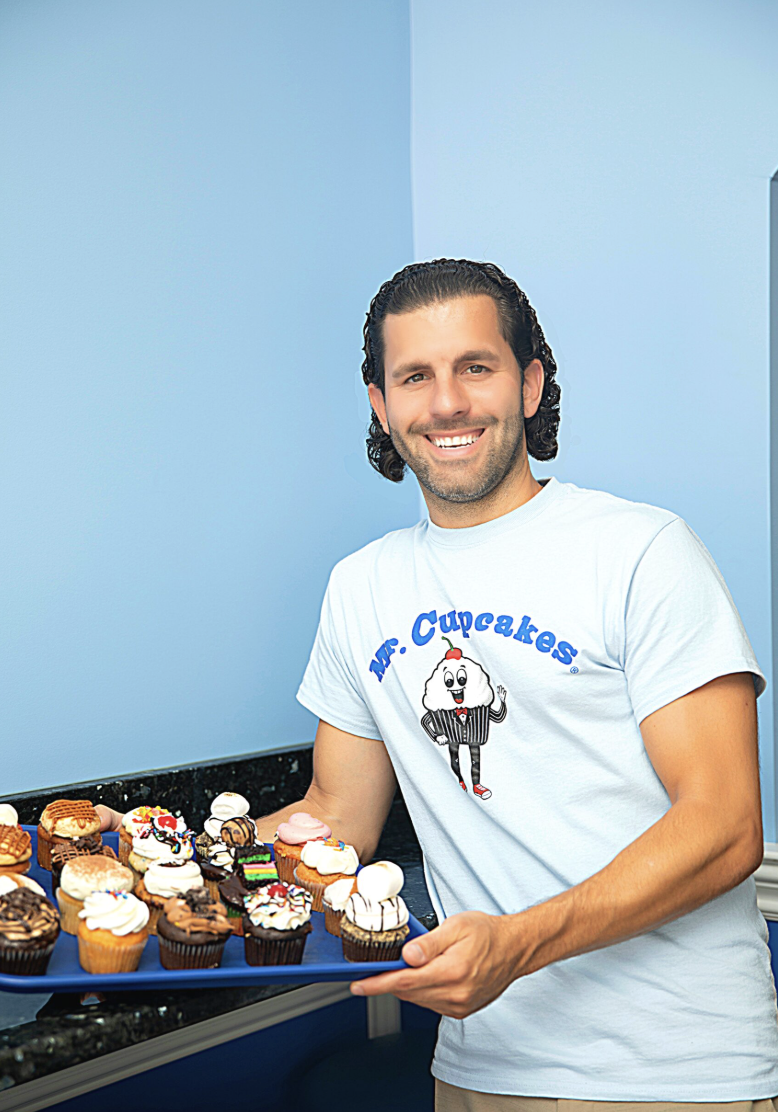 A smiling man holding a tray of various colorful cupcakes in a room with blue walls.