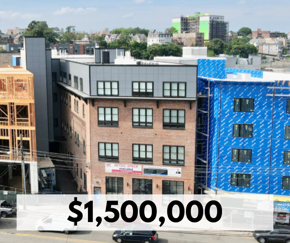 A city street with a new commercial building under construction, a brick building with windows and a retail space for lease, and a blue building wrapped in construction material. Several cars are parked along the street.