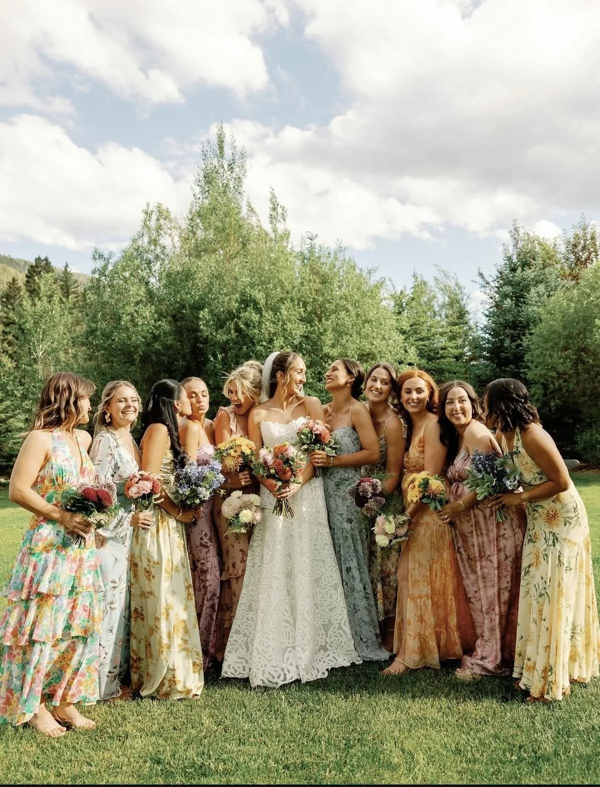 A group of women, including a bride in a white wedding dress, standing outdoors on grass with trees and a cloudy sky in the background, all holding colorful bouquets.