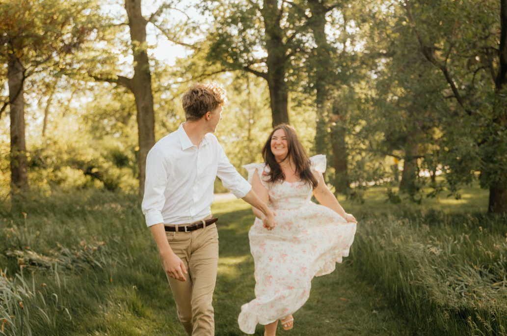A young couple walking through a park in spring or summer, the woman holding her dress while smiling at the man.