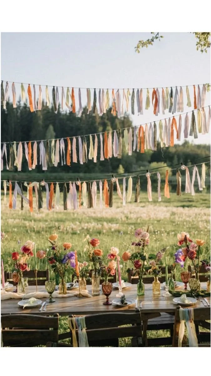 Outdoor festive scene with a long wooden table decorated with colorful flowers in vases, surrounded by chairs with pastel ribbons. String lights and fabric ribbons hang overhead against a backdrop of trees and a field.