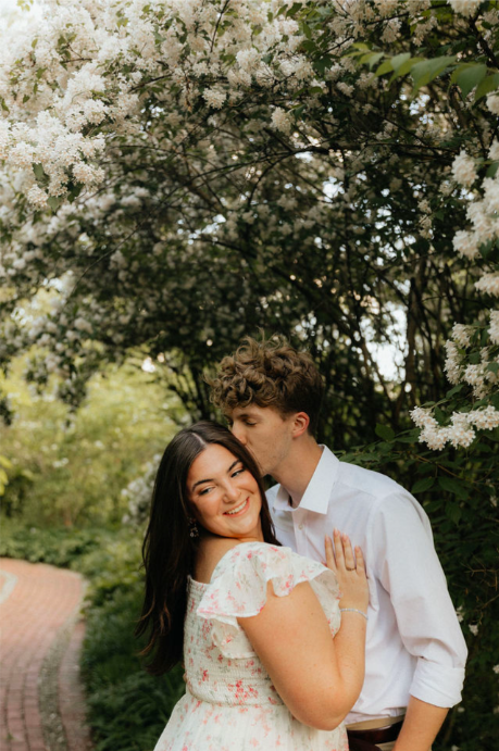 Young couple standing under flowering trees, smiling and embracing outdoors.