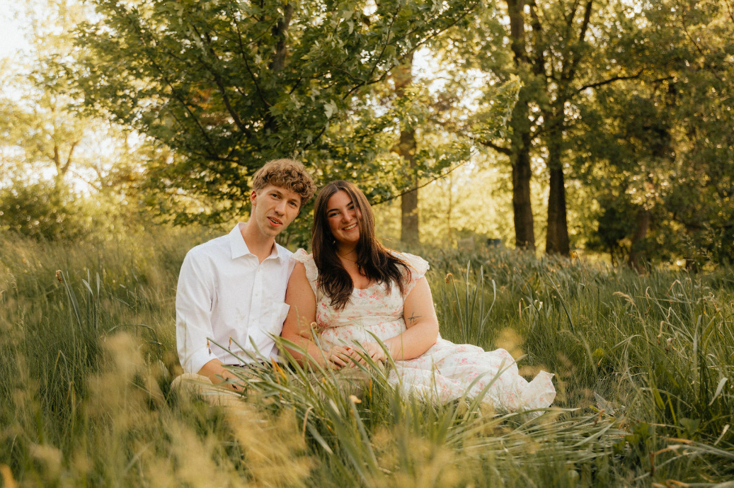 A young couple sitting together on grass in a sunlit forest, smiling for the camera, surrounded by tall grass and trees.
