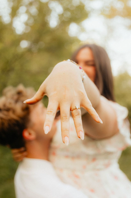A woman showing off a ring on her finger with a person in the background outdoors.
