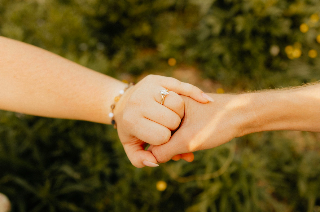 Close-up of two hands clasped, one with an engagement ring, against a green blurred background.