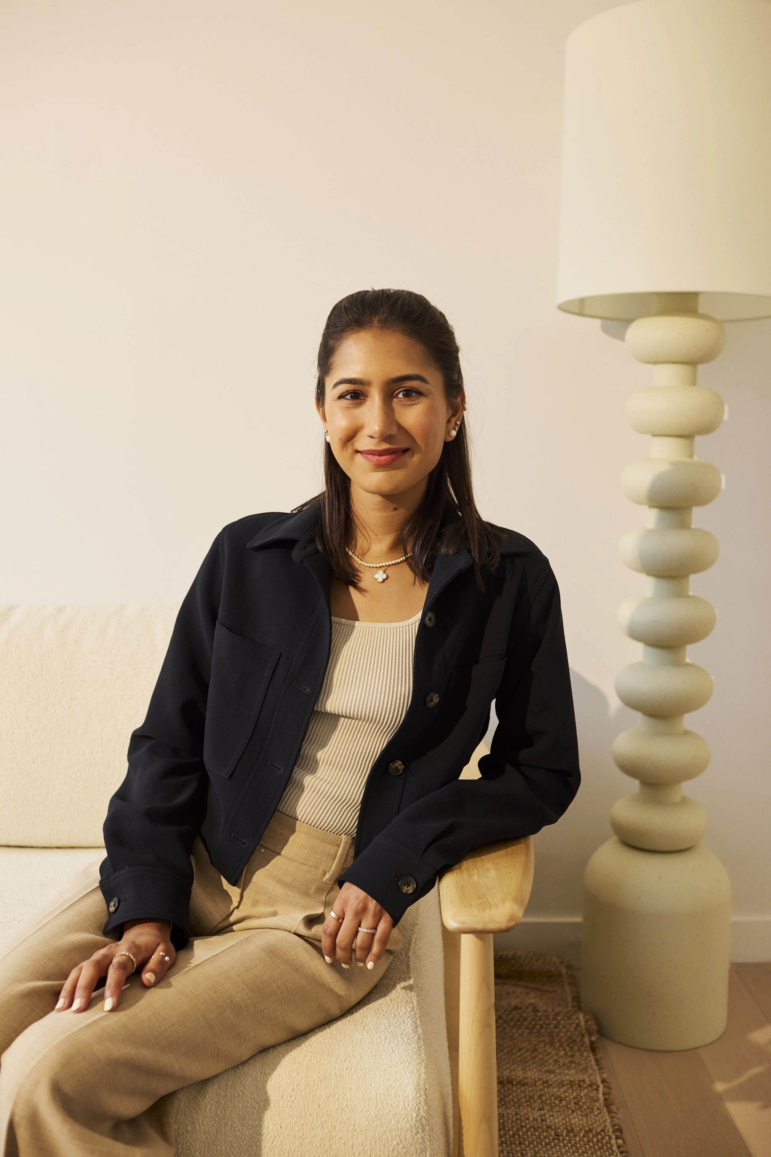 A woman with dark hair, wearing a black jacket, beige top, and beige pants, sitting on a cream-colored couch next to a wooden side table and a tall white lamp in a living room with a neutral decor.