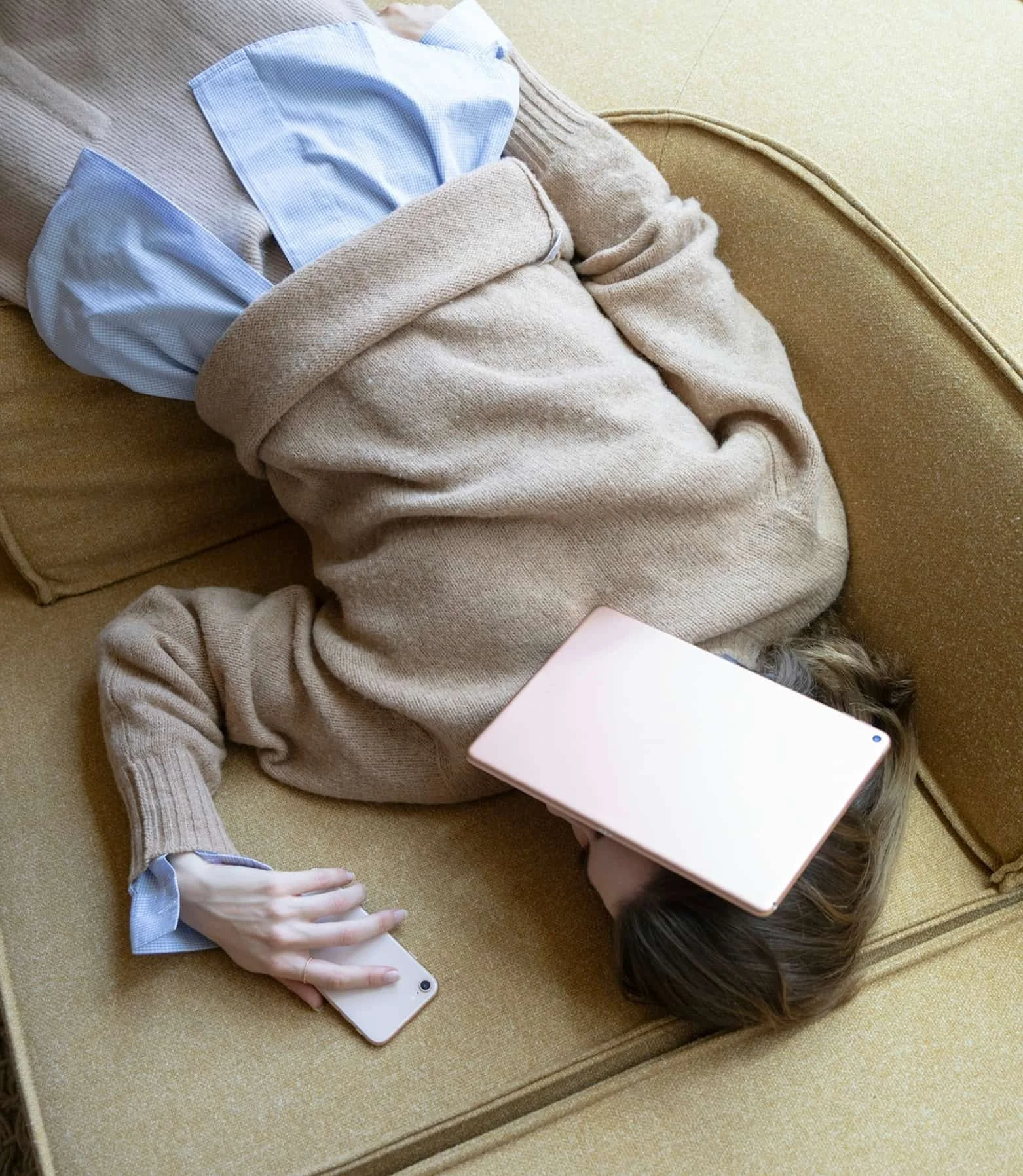 Woman laying on a tan couch with a ipad covering her face and a phone in her hand