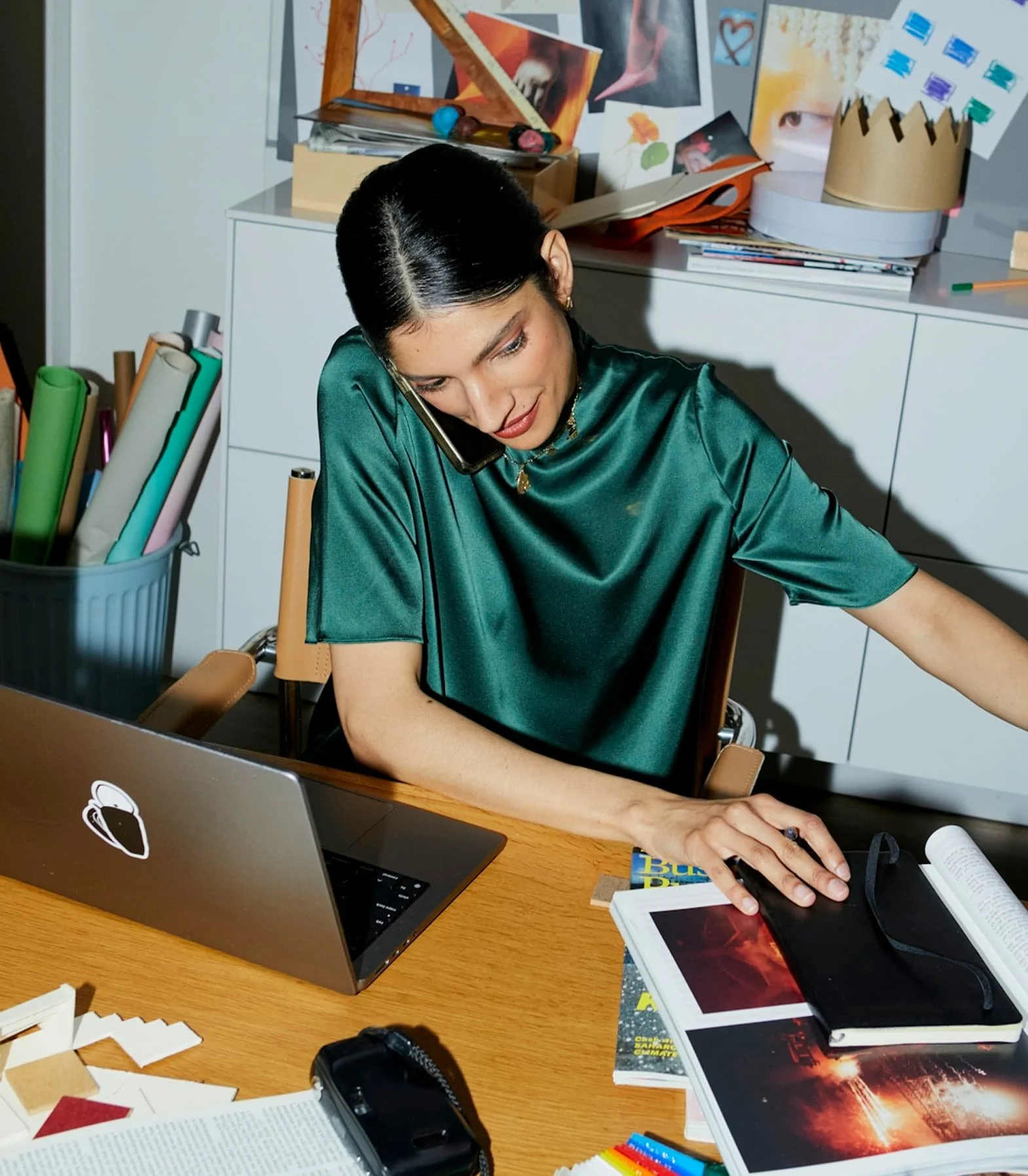 Busy working woman wearing a emerald green shirt. Talking on the phone while on her laptop and sorting though papers and books.