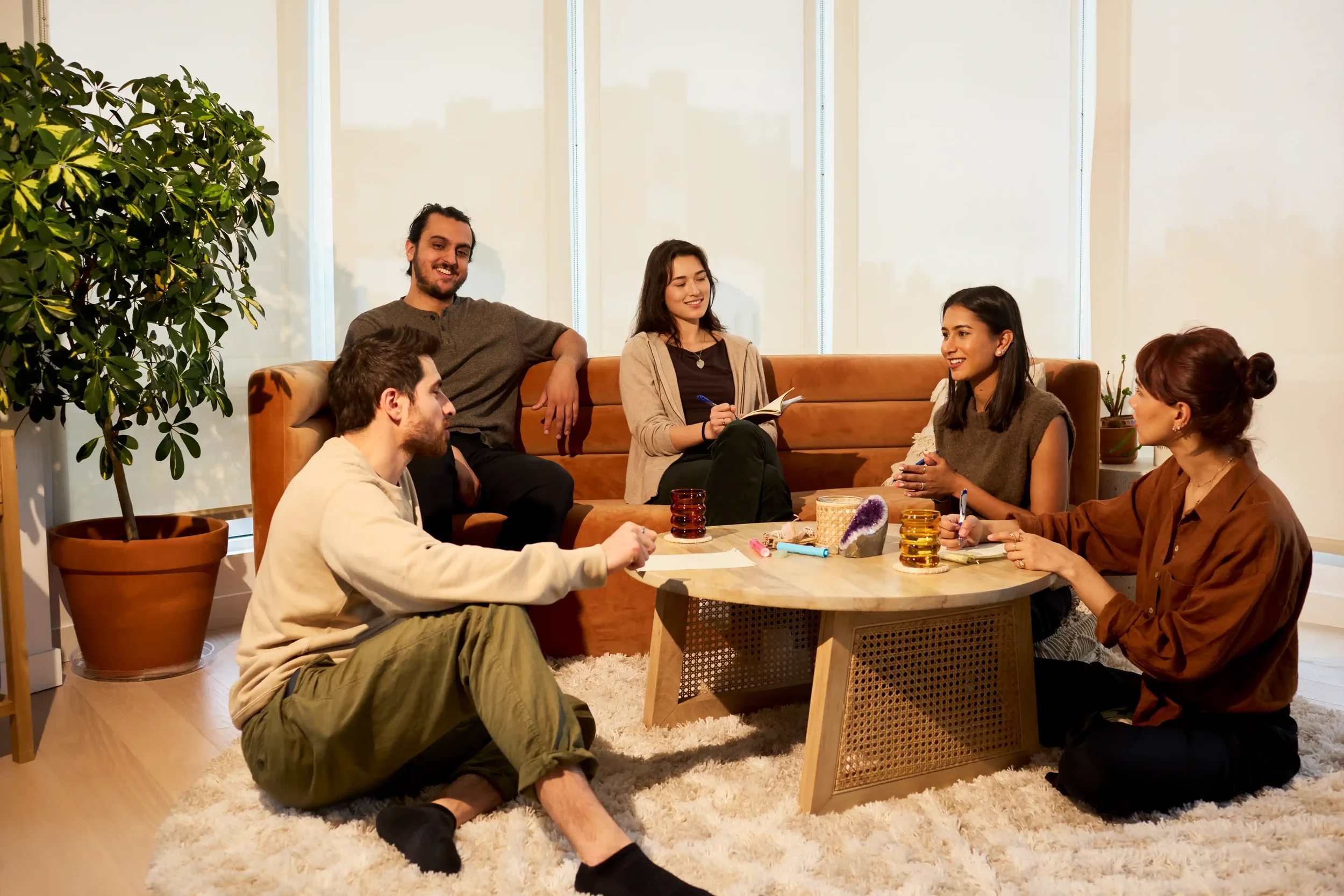 Six people sitting and chatting in a cozy living room with a coffee table and potted plants, enjoying a relaxed gathering.