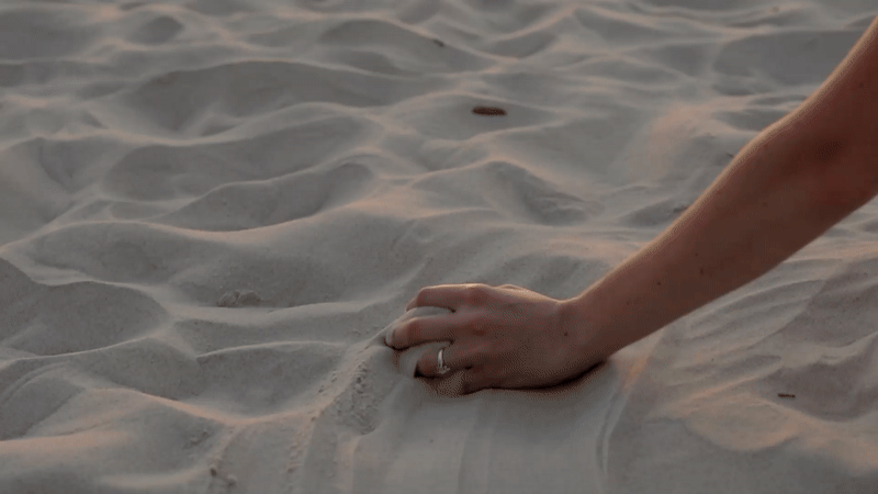 Close-up of a person's hand pressing into white sand on a beach, with a silver ring on the ring finger.