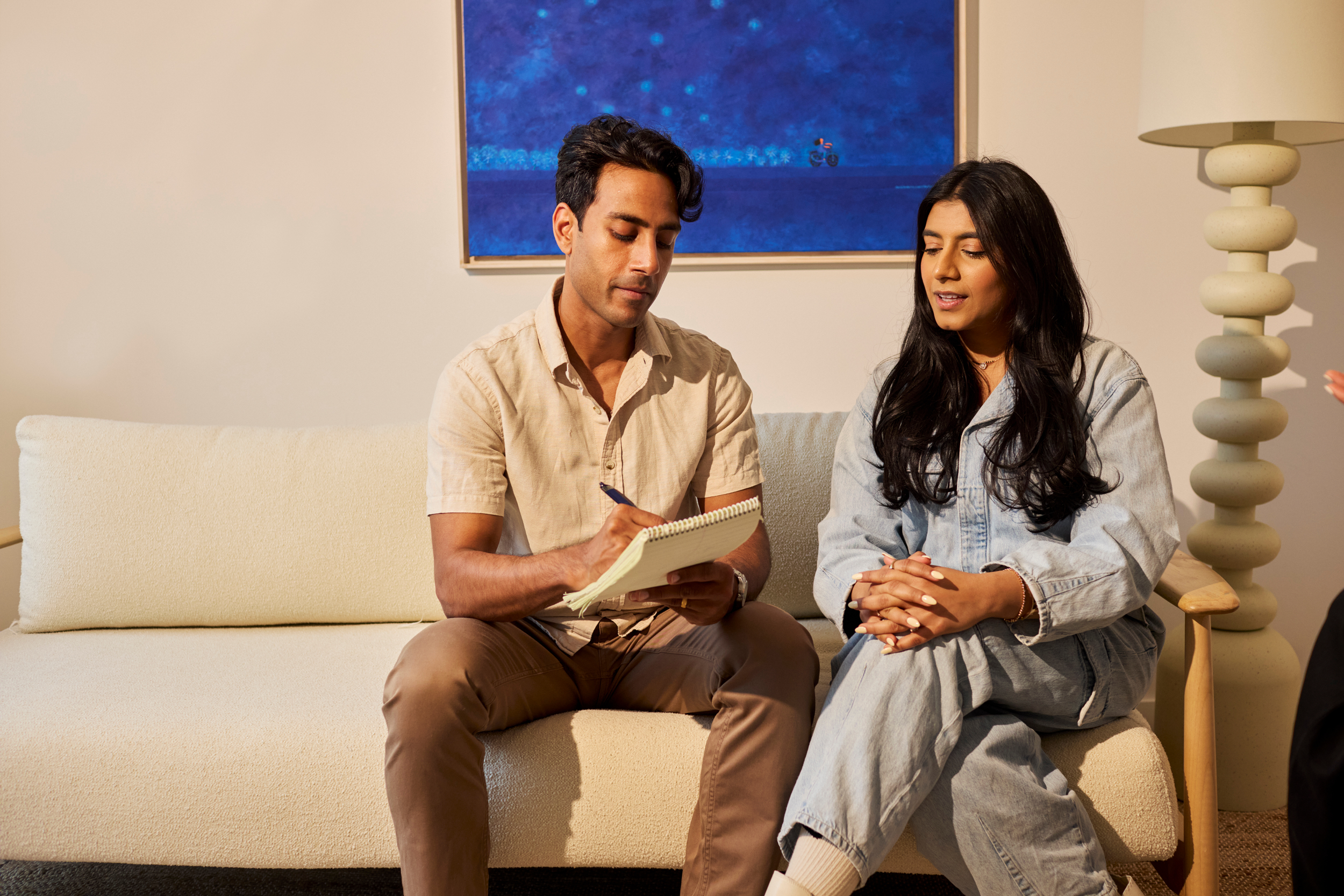 A man and woman sitting on a beige sofa in a therapy or counseling session. The man is taking notes in a notepad, and the woman is speaking with her hands clasped on her lap.