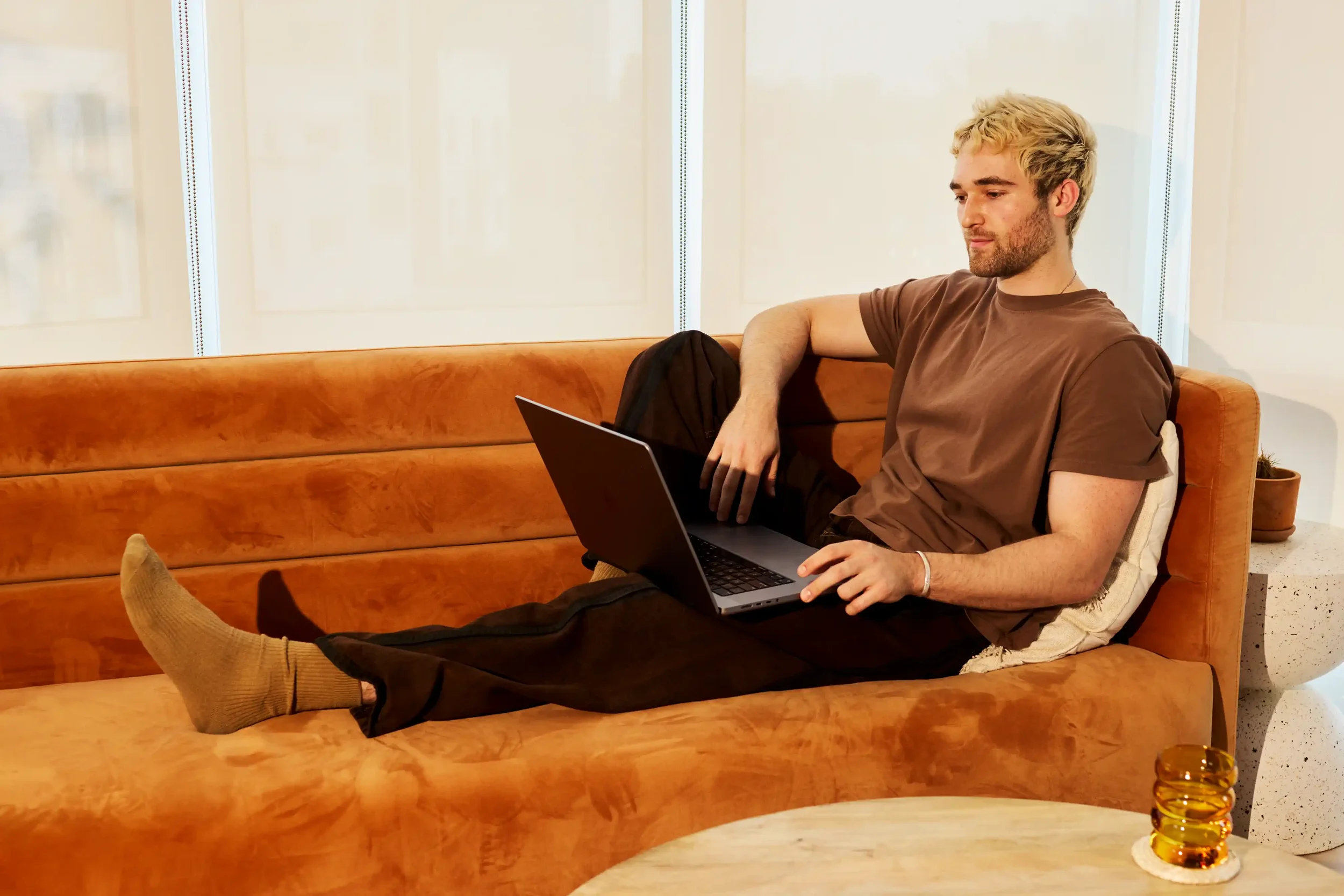 A man with blond hair and a beard relaxing on a brown couch with his legs outstretched, using a laptop, wearing a brown T-shirt, black pants, and socks, in a modern room with large windows and minimal decor.