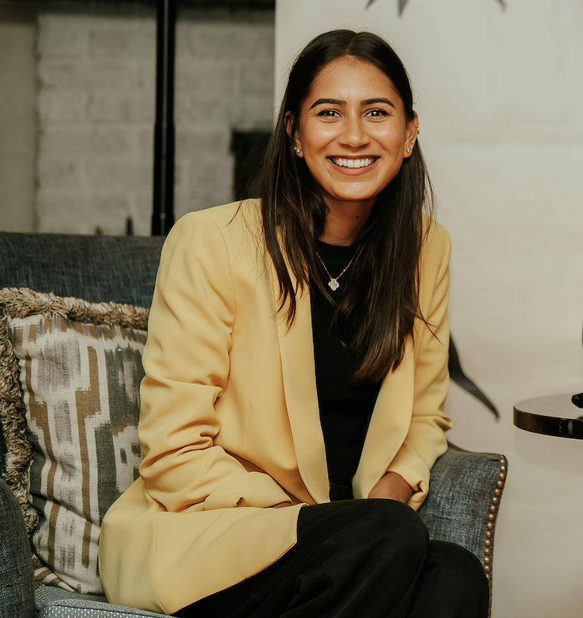 A woman in a yellow blazer sitting on a gray couch, smiling at the camera