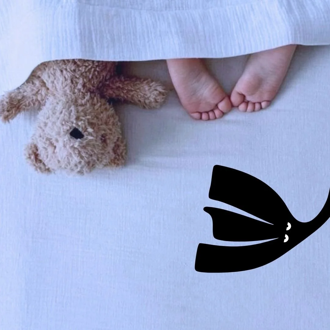 child's feet peeking out of a bed next to a teddy bear and Our Kind Therapy cartoon logo figure