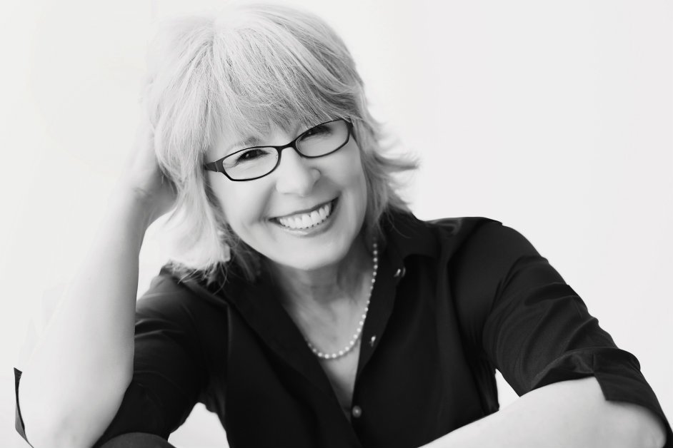 Rebecca LaChance, PhD with short white hair, wearing glasses and a black blouse, posing with her hand touching her head against a plain white background.