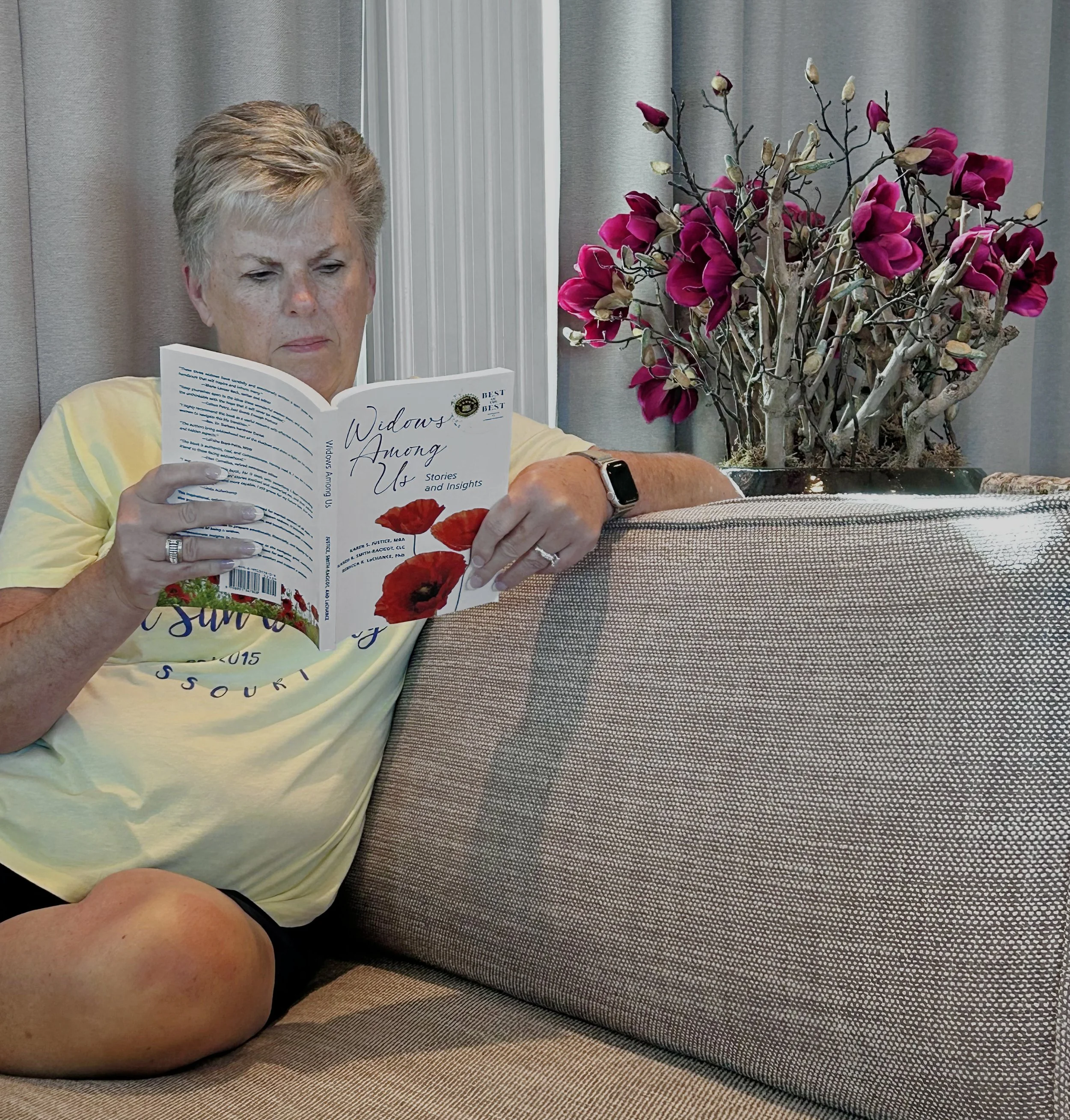 A woman with short blonde hair reading a book titled 'Widows Among Us' sitting on a gray sofa. There is a pink magnolia flower arrangement on a side table next to her, with gray curtains in the background.