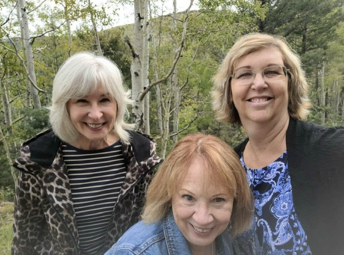 Three women smiling outdoors in a forested area with green trees in the background.