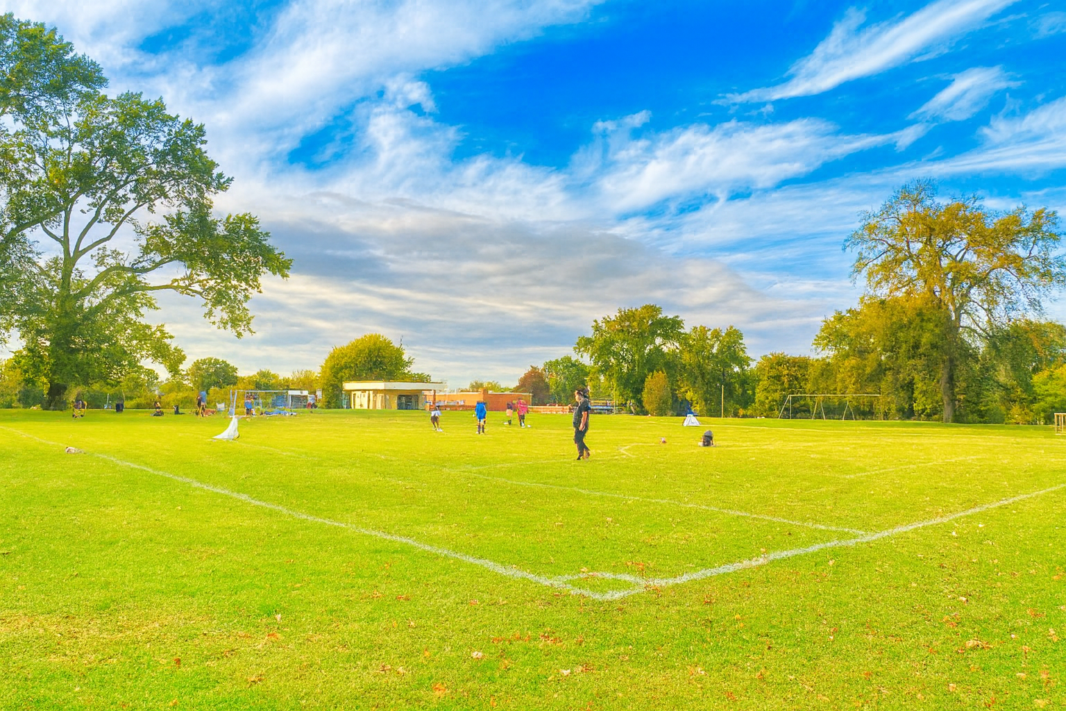 A soccer field with people playing, surrounded by green trees and a partly cloudy sky.