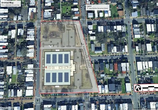 Aerial view of a school campus with two large outdoor solar panel installations in the center, surrounded by a playground and athletic fields, located in a residential neighborhood.