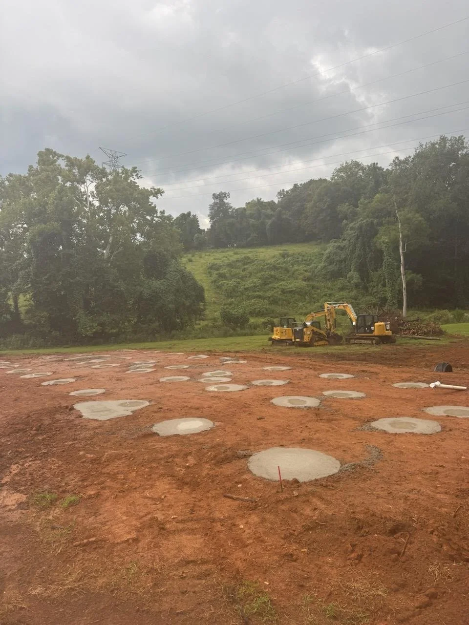 Construction site with round concrete pads on reddish dirt ground, excavators in the background, and natural landscape with trees and a cloudy sky.