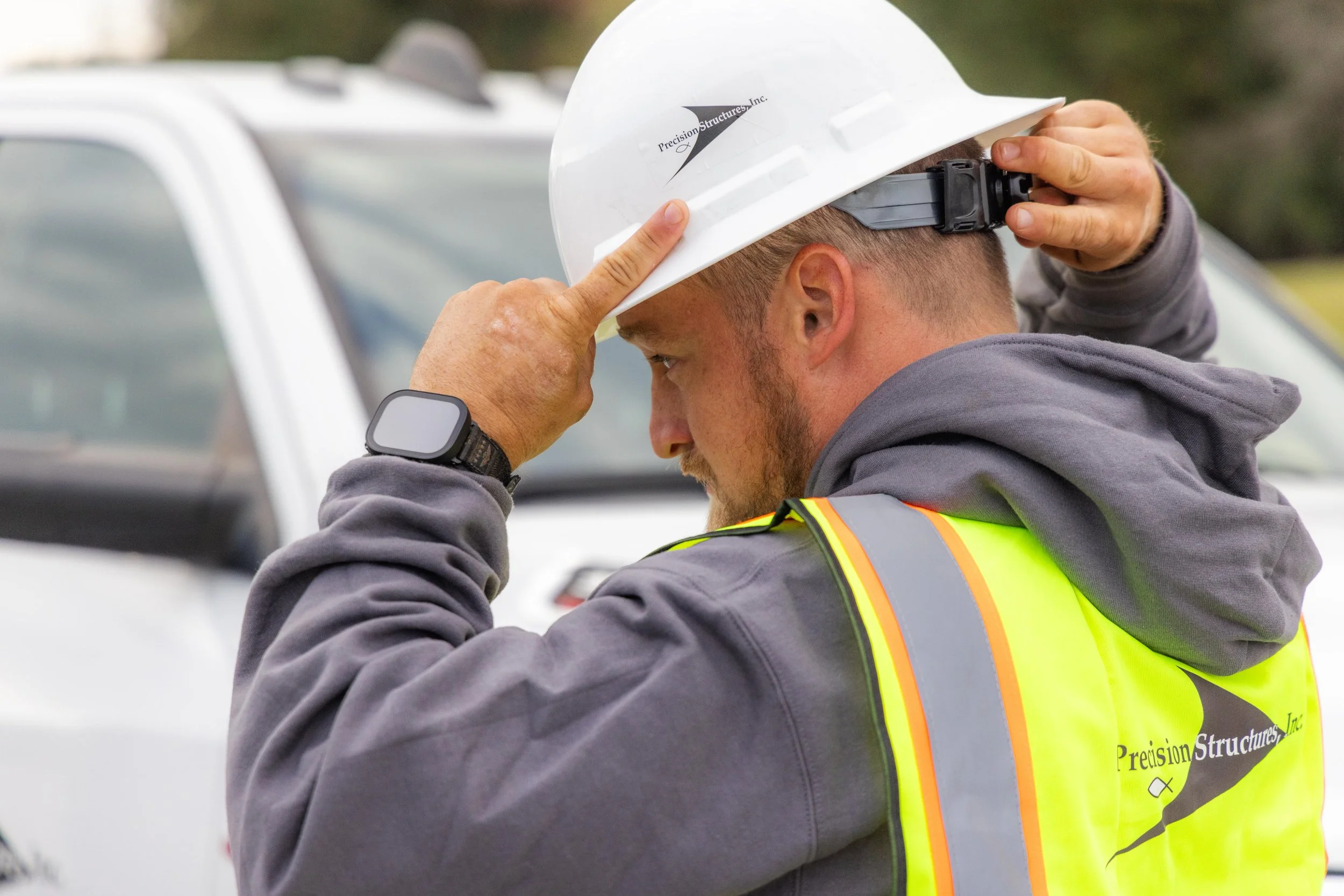 A construction worker wearing a white safety helmet and a yellow-green reflective vest adjusting his helmet.